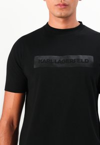 Svart t-skjorte med kort erme laget av bomull, med en matt svart rektangulært logo hvor "KARL LAGERFELD" er trykket med fet skrift.