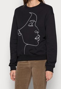 Sudadera negra con un diseño de cara dibujada a mano en blanco, puños y dobladillo acanalados, con cuello redondo y corte relajado.