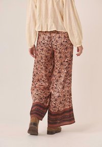 Pantalones fluidos y estampados en tonos cálidos presentan diseños florales y un dobladillo de patrón contrastante. Se combinan con una blusa de color claro y texturizada.