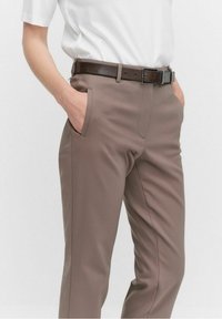 Osoba ubrana w spodnie taupe z bocznymi kieszeniami i ciemnobrązowy skórzany pasek, ręce częściowo w kieszeniach, biała koszula z krótkim rękawem.