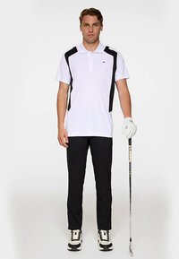 Vit pikétröja med svarta sidopaneler, svarta byxor och svartvita golfs skor. Håller i en golfklubba, bär en golfhandske.