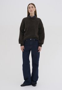 Brauner Strickpullover mit hohem Kragen, kombiniert mit dunkelblauen weit geschnittenen Jeans, schwarzen Schuhen und einem darunter sichtbaren Hemd mit Kragen.