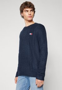 Pull en maille bleu marine avec col rond, texture en cable, et un petit patch logo rouge, blanc et bleu en haut à gauche.