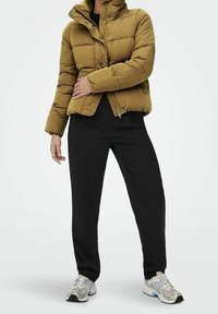 Brązowa kurtka puchowa z wysokim kołnierzem, charakteryzująca się pikowanymi sekcjami, w połączeniu z czarnymi spodniami i szarymi trampkami z niebieskimi akcentami.