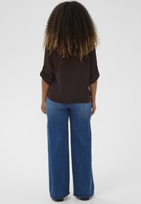 Pruun, lahtine pluus, mille varrukad on üles rullitud, yhdatud laiade siniste teksadega. Kangas paistab pehme ja sileda tekstuuriga.