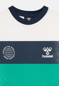 Közelkép egy póló nyakáról, tengerészkék bordázott szegéllyel és vízszintes színblokkokkal fehérben, tengerészkékben és zöldeskékben, tengerészkék szalagon logókkal és szöveggel.