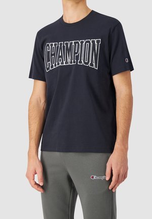 Mężczyzna ubrany w granatowy T-shirt z napisem „CHAMPION” oraz szare spodnie dresowe z małym logo Champion na udzie, stojący na jednolitym tle.