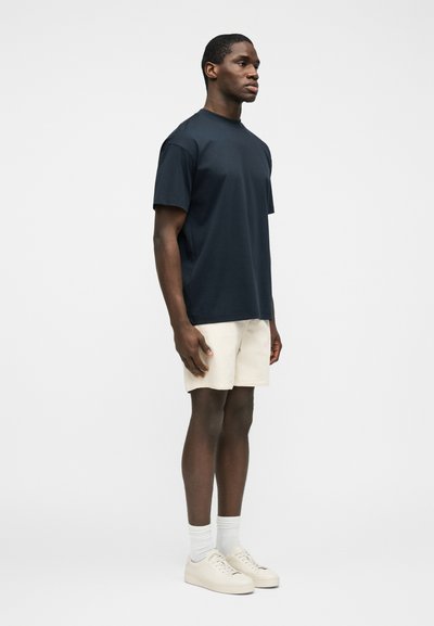 Άνδρας που στέκεται φορώντας μπλε σκούρο μπλουζάκι, μπεζ σορτς, λευκές κάλτσες και λευκά αθλητικά παπούτσια μπροστά σε απλό ανοιχτό γκρι φόντο.