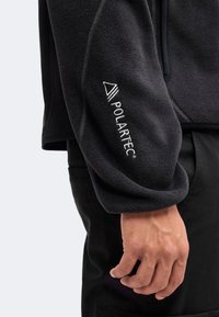 Κοντινό πλάνο σε μανίκι μαύρου fleece μπουφάν με το λευκό λογότυπο Polartec κεντημένο κοντά στον καρπό, φορεμένο από άτομο με χαλαρό χέρι.