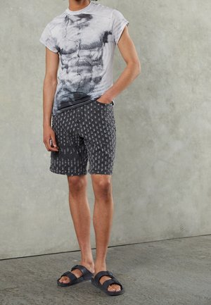 Graues T-Shirt mit einem rissigen Textur-Graphic, kombiniert mit schwarzen gemusterten Shorts und schwarzen Slide-Sandalen. Lässiges Outfit vor einem neutralen Hintergrund.