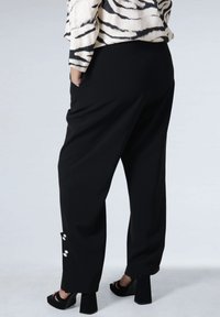 Woman wearing black wide-leg pants with silver buttons near the hem, black heeled sandals, and a cream zebra-patterned top.