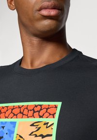 Black cotton t-shirt features a graphic design with rectangular blocks in orange, blue, and yellow, outlined with a bright green border.