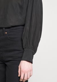 Svart blus med en mjuk textur och voluminösa ärmar, i kombination med svarta jeans som har en kopparknappdetalj.