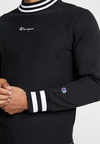 Felpa nera con colletto alto, caratterizzata da strisce bianche. Logo Champion sul petto, polsini a costine con strisce bianche, texture morbida.