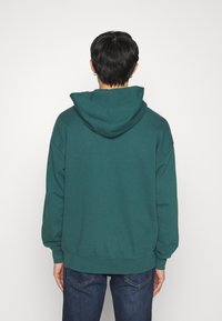 Turkusowa bluza z dużym, regulowanym kapturem, prążkowanymi mankietami i luźnym krojem, w zestawieniu z ciemnoniebieskimi dżinsami. Gładka tekstura materiału.