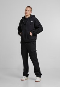 Un sweat à capuche noir zippé avec des poches kangourou, associé à un pantalon cargo noir. La tenue présente une coupe décontractée et des baskets blanches avec des accents rouges.