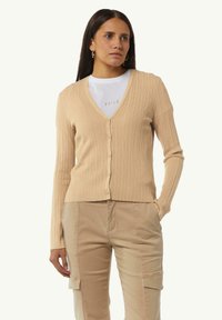 Beige gerippter Cardigan mit V-Ausschnitt, langen Ärmeln und fünf Knöpfen auf der Vorderseite. Getragen über einem weißen T-Shirt mit aufgedrucktem Text.
