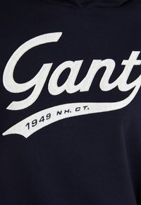 Navy blue sweatshirt with white embroidered "Gant" logo and "1949 NH CT" text on front. Smooth texture, casual fit.