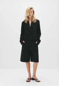 Čierny skrátený blazer s jedným gombíkom, skombinovaný s voľnými čiernymi šortkami. Obe majú hladkú textúru a minimalistický dizajn. Čierne sandále na remienky.