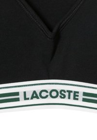 Top negro de cuello en V con textura acanalada. La banda blanca en el dobladillo presenta el texto "LACOSTE" en verde y rayas.
