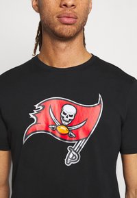 T-shirt en coton noir avec un grand design de drapeau rouge comportant un crâne, des épées croisées et un ballon de football. Encolure ras du cou simple.