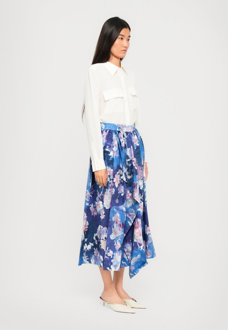 Donna in una blusa bianca con tasche frontali e una gonna midi floreale blu abbinata a décolleté bianchi con punta.