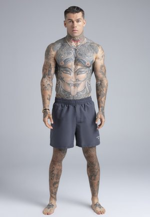 Lihaseline mees, kellel on laialdased tätoveeringud, sealhulgas suur nägu torso peal, seljas tumedad lühikesed püksid, paljajalu seistes ühetaolise tausta ees.