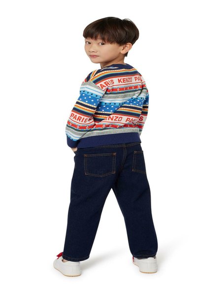 Pull en tricot coloré avec des rayures horizontales, avec du texte en rouge et bleu. Équilibré avec un pantalon en denim bleu foncé et des baskets blanches.