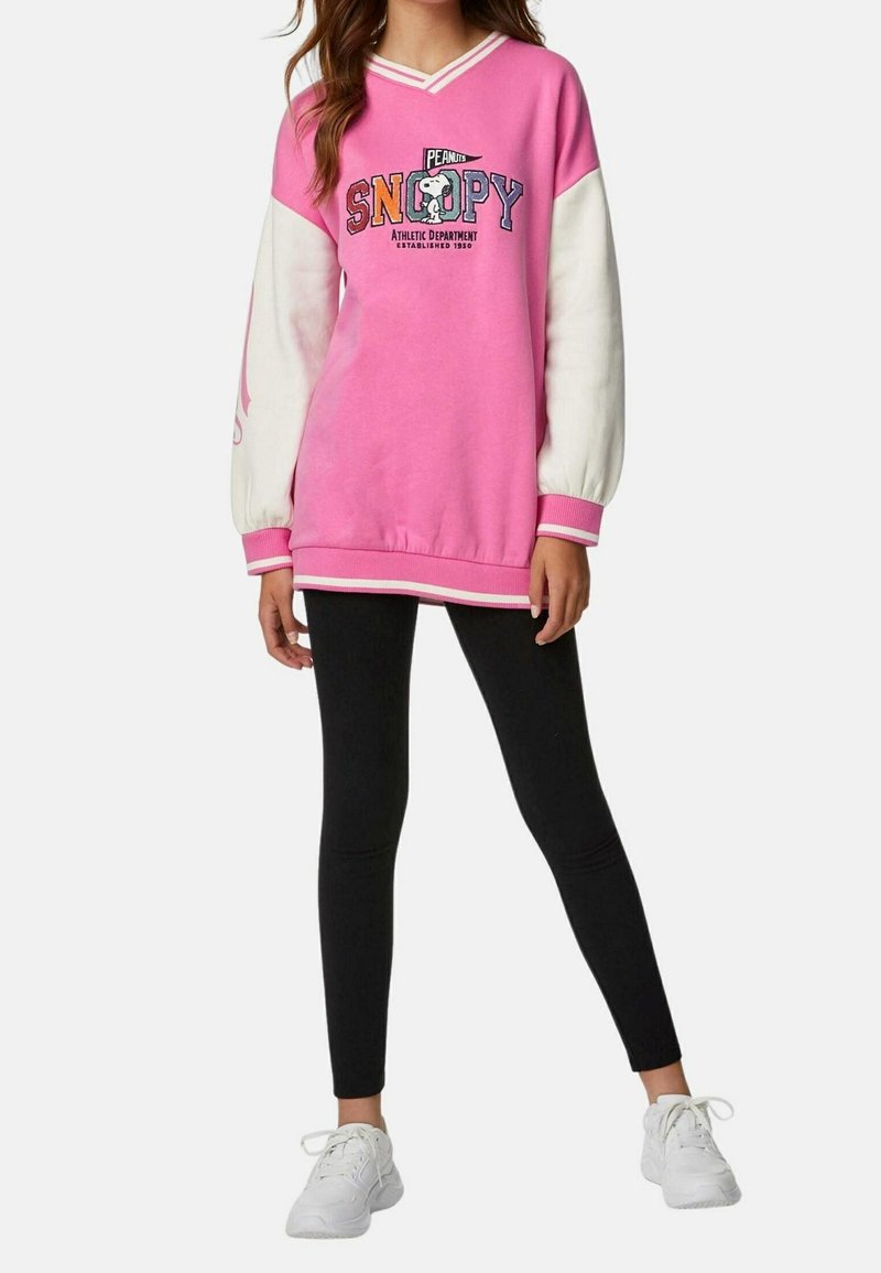 Młoda osoba w różowo-białej bluzie z Snoopy, czarnych legginsach i białych trampkach stoi na jednolitym tle.