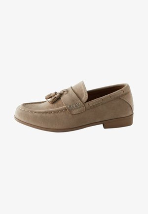 Beige Wildleder Slipper mit einer strukturierten Oberseite, dekorativen Quasten und subtilen Nähten im oberen Bereich. Niedriger Gummiabsatz für zusätzlichen Komfort.