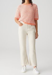 Mujer con un top de punto de rayas coral y blanco, pantalones de cintura alta acampanados en color blanco roto y zapatillas blancas, de pie con una mano en el bolsillo.