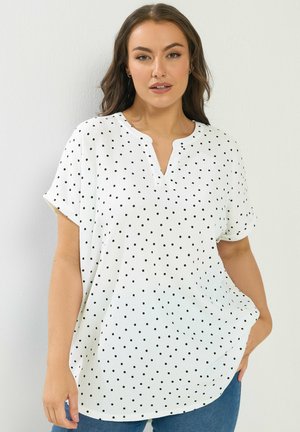 Kobieta ubrana w białą bluzkę z krótkim rękawem w czarne kropki, z dekoltem w kształcie litery V, oraz niebieskie dżinsy, pozująca na tle jednolitego jasnego tła.