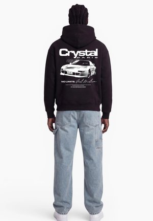 Person wearing a black hoodie with a white sports car and "Crystal Paris" text on the back, paired with light blue jeans and white shoes.