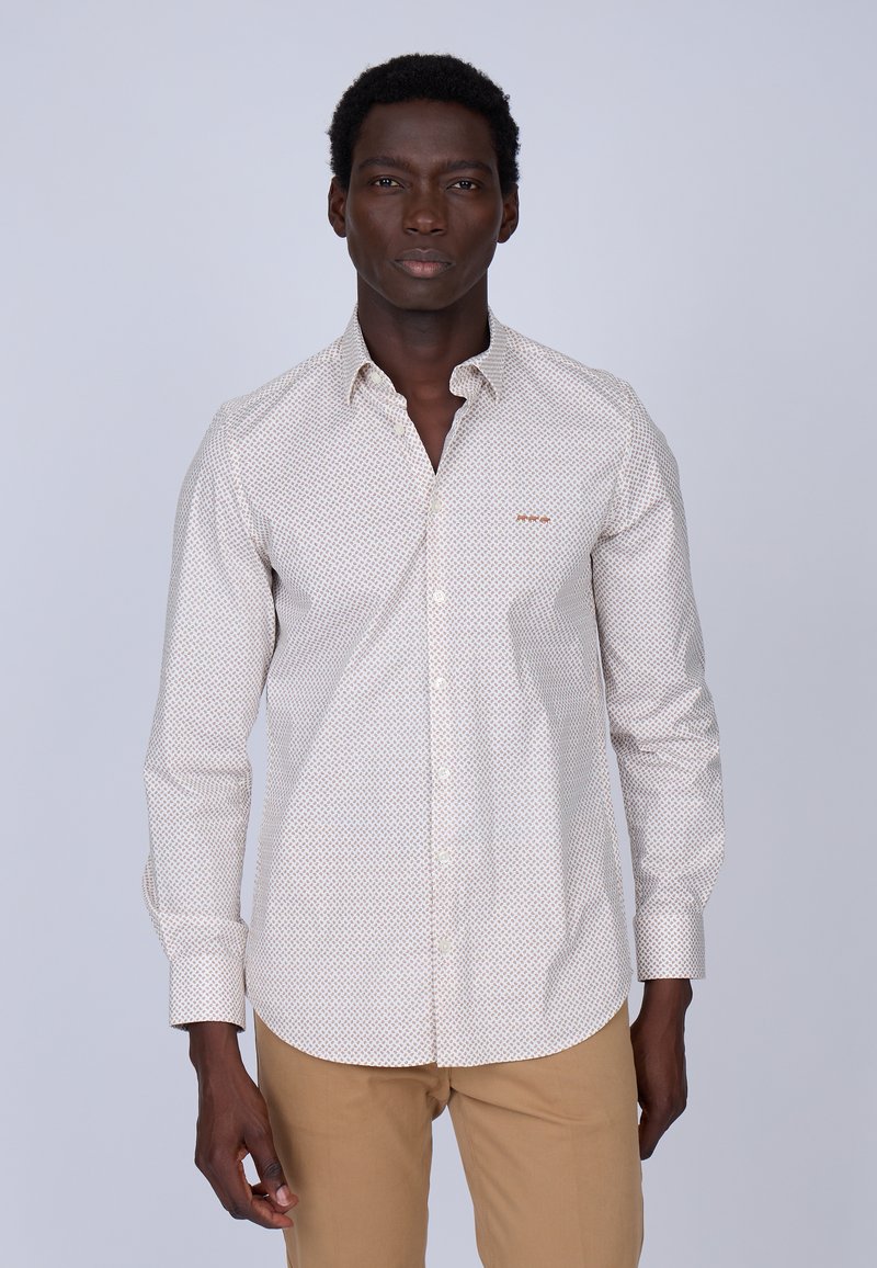 Chemise boutonnée de couleur claire avec un motif géométrique, à manches longues et avec un logo sur la poitrine. Portée avec un pantalon beige, présentée sur un fond neutre.