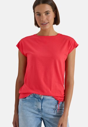 Jeune femme portant un haut rouge à manches courtes et un jean bleu clair, debout devant un fond blanc uni.