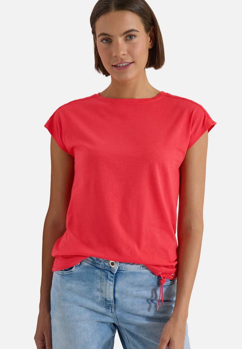 Jeune femme portant un haut rouge à manches courtes et un jean bleu clair, debout devant un fond blanc uni.