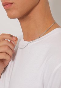 Sølv kæde vedholdt af en hånd, båret om en bar hals. Kæden har en glat, skinnende overflade og et fleksibelt design. Hvid skjorte nedenunder.
