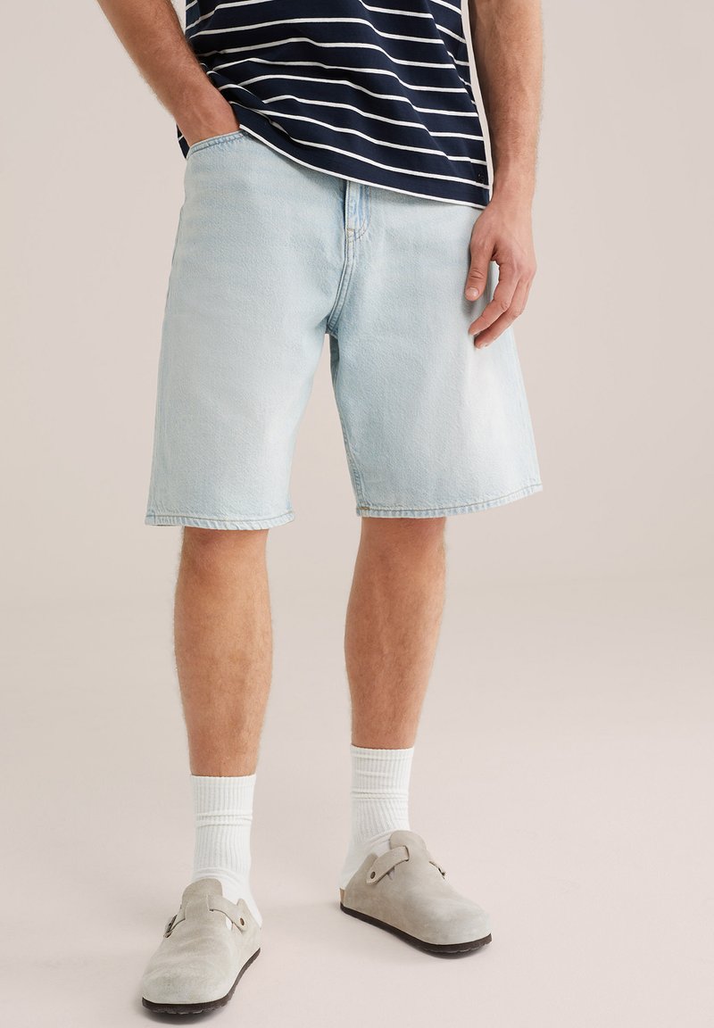 Mees, kes kannab helesiniseid teksapükse, valgeid sukki, halli libisevaid jalatseid ja tumesinise-valge triibulist särki.