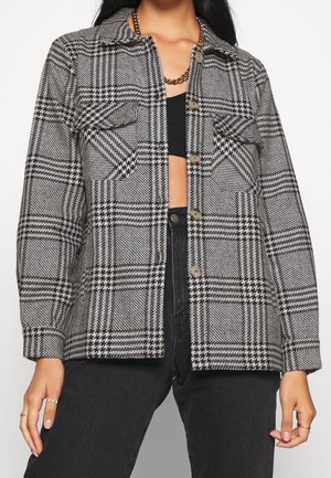 Femme portant une veste boutonnée à carreaux noir et blanc par-dessus un crop top noir et un jean foncé, avec un collier chaîne en or.