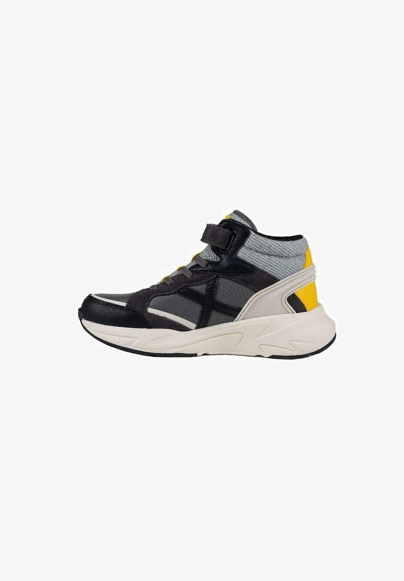 Zapatilla deportiva alta con parte superior negra, acentos de malla gris, detalle en el talón amarillo y suela beige. Cuenta con una correa de Velcro para un ajuste seguro.