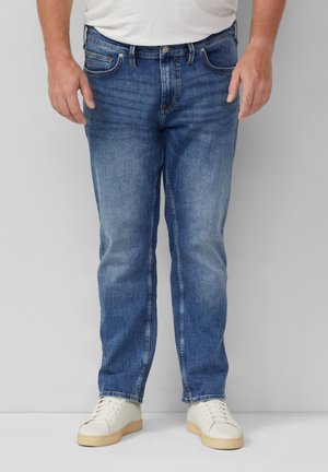 Man draagt mediumblauwe rechtvallende jeans, witte sneakers en een wit shirt, staand tegen een eenvoudige grijze achtergrond.