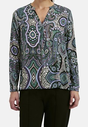 Frau trägt eine langärmlige Bluse mit Paisley-Muster in Grün, Schwarz, Lila und Weiß, kombiniert mit schwarzen Hosen, steht mit entspannten Armen.