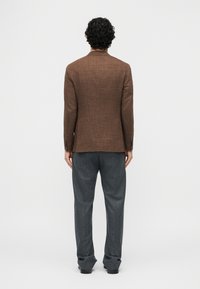 Άνδρας με σγουρά μαλλιά, φοράει εφαρμοστή καφέ σακάκι και ανοιχτό γκρι παντελόνι, στέκεται με την πλάτη προς το μέρος μας μπροστά σε απλό λευκό φόντο.