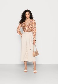 Blusa floral em tons de ferrugem e creme com babados; calças beje de cintura alta; mala de ombro neutra; sandálias brancas de plataforma trançadas.