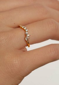 Guld ring med bølge-design, prydet med små, klare ædelsten placeret langs den øverste kant. Båret på en tekstureret hånd.