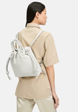 Weißer Leder-Zugbeutel-Rucksack mit geknoteten Griffen, glatter Textur und verstellbaren Riemen, getragen auf dem Rücken einer Person in einem beigen Hemd.