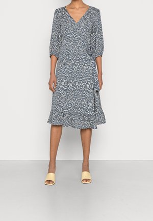 Femme portant une robe midi bleue à imprimé floral blanc, col en V, manches trois-quarts, ourlet à volants, et sandales jaunes ouvertes à l'avant.