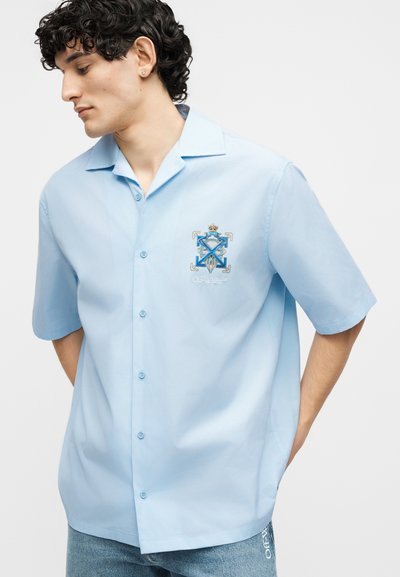 Νεαρός άνδρας που φοράει ανοιχτό μπλε κοντομάνικο πουκάμισο με κουμπιά και κεντημένο μπλε και χρυσό έμβλημα στο στήθος, σε συνδυασμό με ανοιχτόχρωμα τζιν.