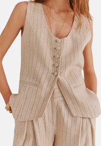 Beige Leinenweste mit vertikalen Streifen, tiefem V-Ausschnitt, Front mit fünf Knöpfen und aufgenähten Taschen. Der Stoff hat eine strukturierte Oberfläche.