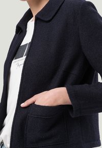 Navy wool jacket with a collar, buttonless front, two pockets, and a textured fabric. Worn over a white shirt with a graphic design.